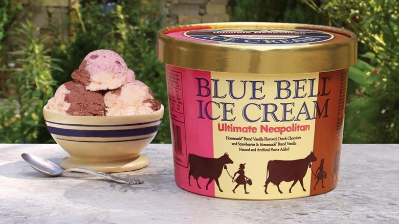 A carton of Blue Bell Ultimate Neapolitan ice cream on an outdoor patio table next to a bowl of ice cream