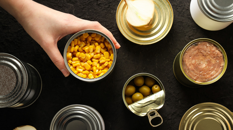 Top view of hand reaching for canned corn among various canned foods