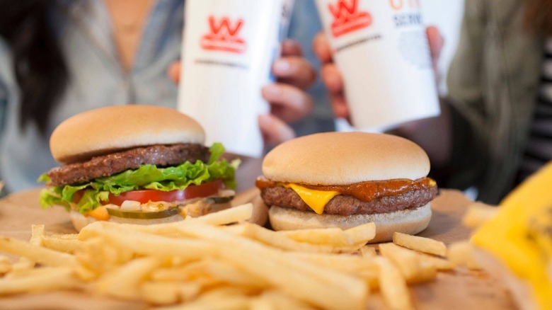 A regular burger sitting beside a chili cheeseburger from Wienerschnitzel with fries in the foreground and two people with fountain drinks in the background.