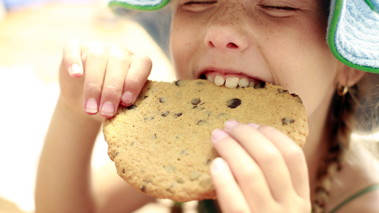 A child takes a bite of a large chocolate chip cookie
