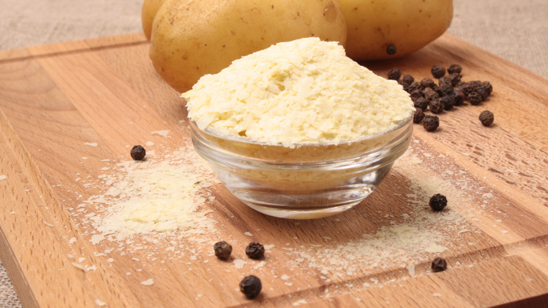 Instant potato flakes in a glass bowl on a wood cutting board surrounded by whole potatoes and peppercorns