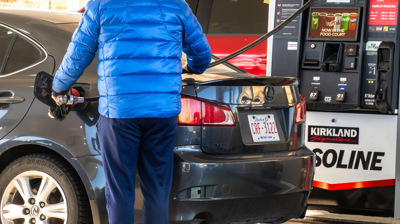 A person in a blue jacket is seen from behind, filling their gray car with Kirkland Signature gasoline at a fuel pump