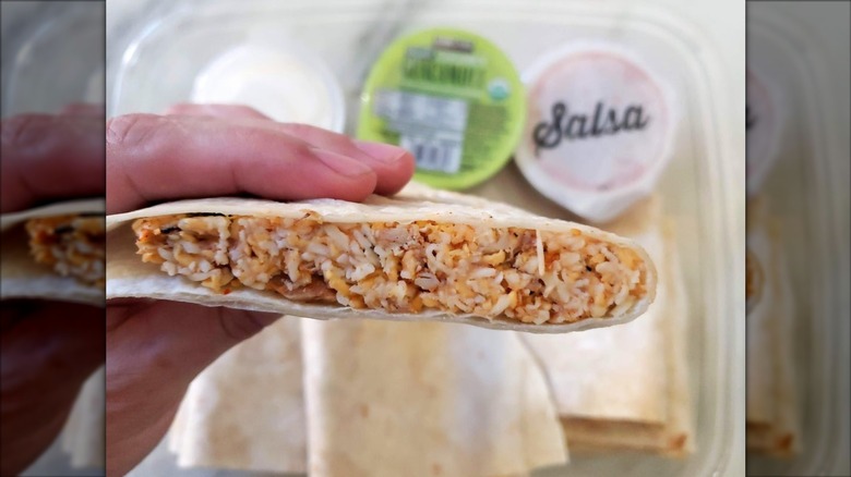 A close-up image of a person holding a prepared Kirkland Signature Chicken Quesadilla with the packaging and sauces in the background