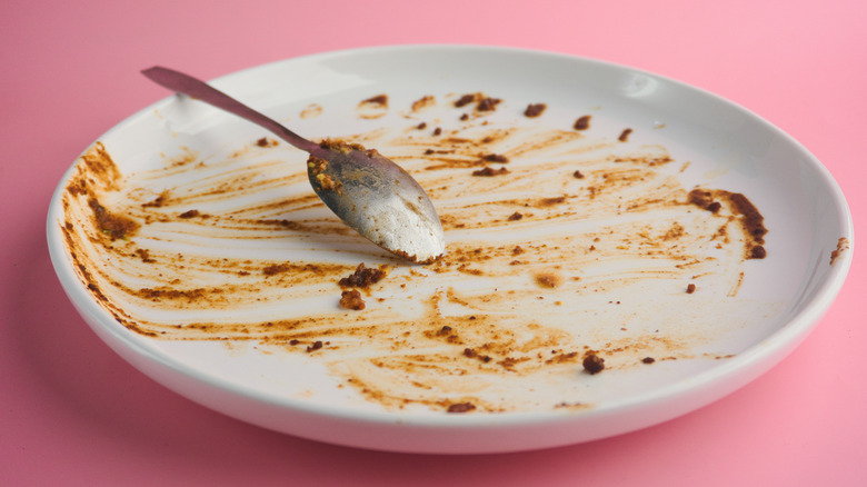 Close-up of an empty white plate with food remnants and a spoon against a pink background