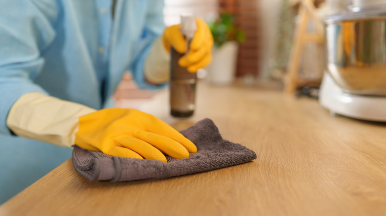 A woman wearing yellow gloves cleaning a wooden countertop with disinfectant spray and a cloth