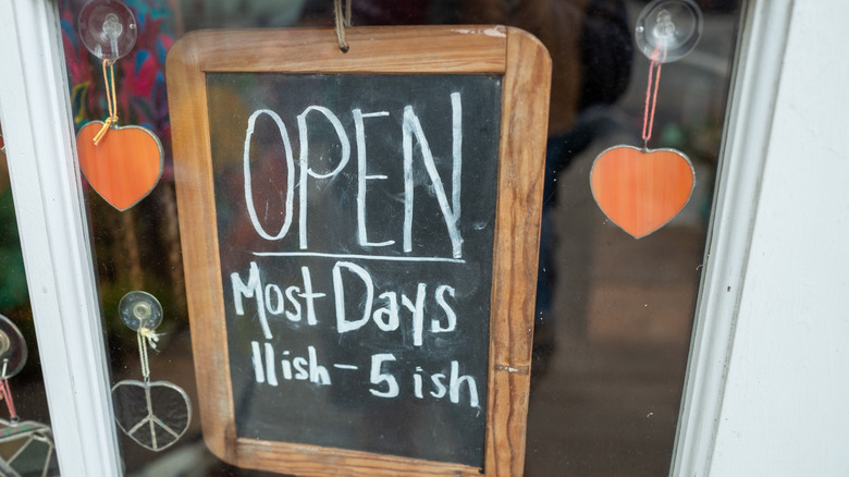 A chalkboard sign hanging in a store window that says open most days 11ish to 5ish