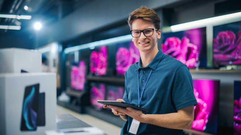 A man holding a tablet smiles in front of a wall of televisions
