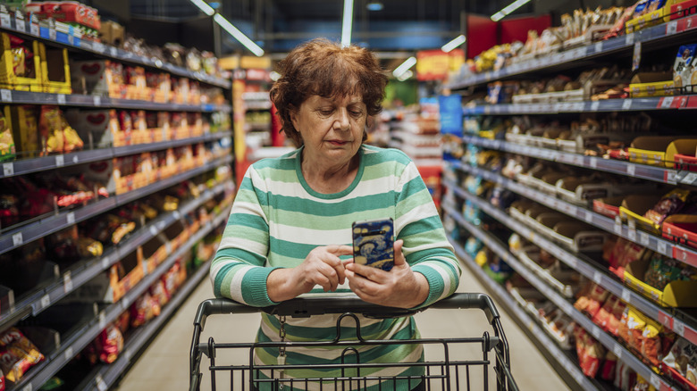 A woman standing in a supermarket aisle looking at her cellphone with a shopping cart