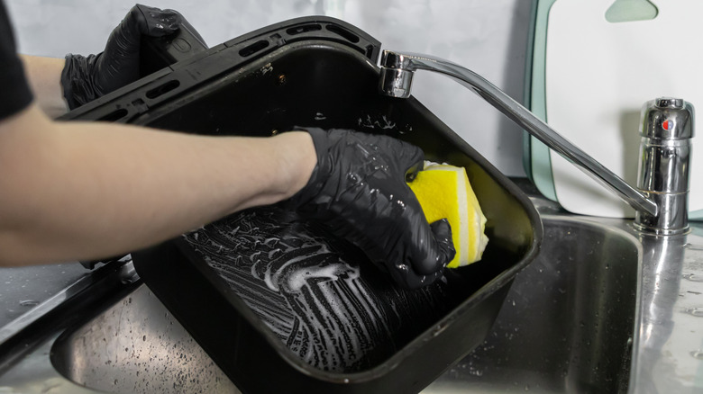 A person wearing black gloves using a yellow sponge to clean an air fryer basket in a sink
