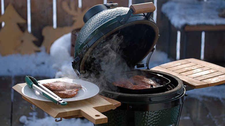 A small charcoal grill in the snow with the lid open and cuts of meat on the grill and on a plate nearby