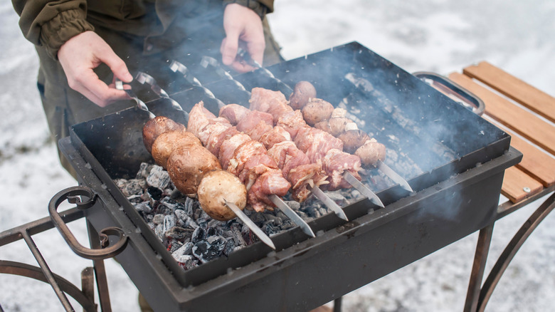 A person tending to skewers of meat over a charcoal grill in the snow