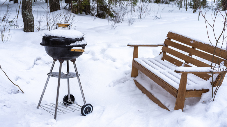 A small charcoal barbecue grill covered in snow next to a snowy wooden bench