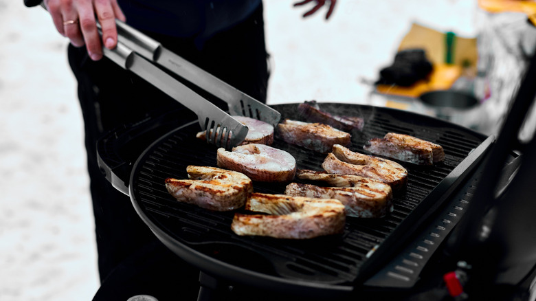 A close-up of a person using tongs to turn meat while grilling outdoors in the snow on a charcoal grill