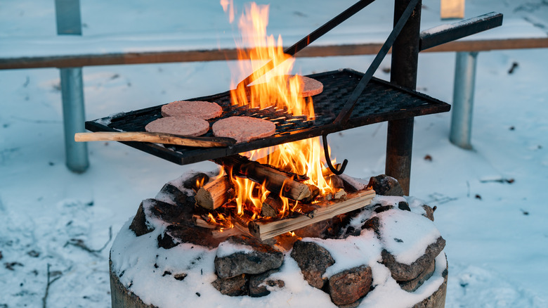 Hamburger patties cooking outdoors in the snow on a cast iron grill over a wood-burning fire