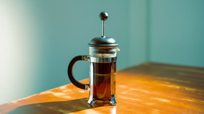A sunlit French press coffee maker on a wooden table with a teal background.