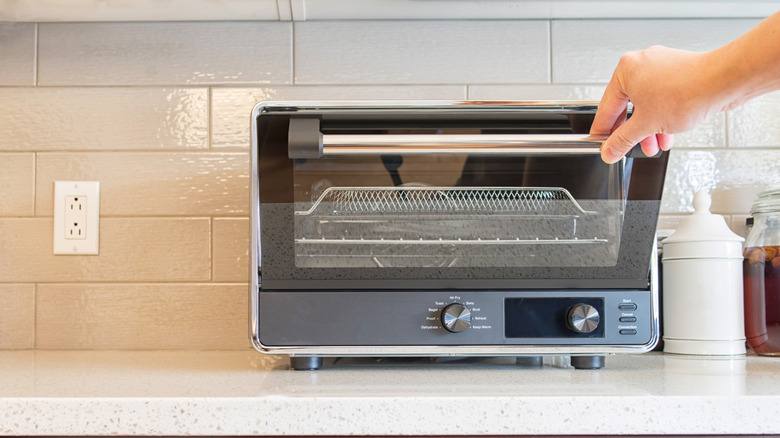 Hand opening a gray toaster oven sitting against white tile backsplash