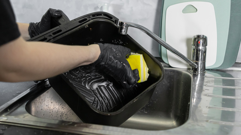 A gloved hand scrubbing an air fryer basket with a soapy yellow sponge