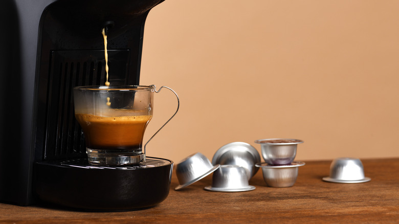 Black single-serve coffee maker brewing coffee in glass cup, next to silver capsules