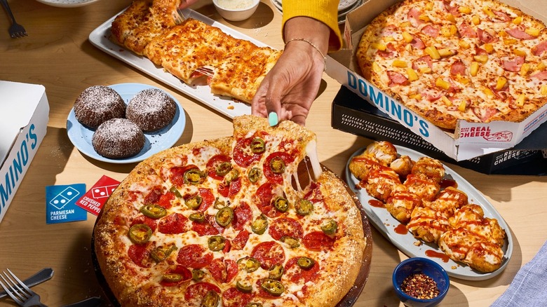a hand grabbing a slice of pizza from a table laden with pizzas, breadsticks, lava cakes, and other Domino's offerings