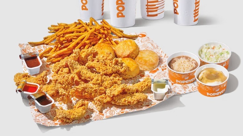 Popeyes chicken tenders, fries, biscuits, and dipping sauces laid out beside containers of side dishes and drinks