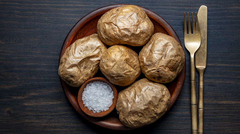 Plain baked potatoes in wooden bowl with bowl of salt, next to cutlery