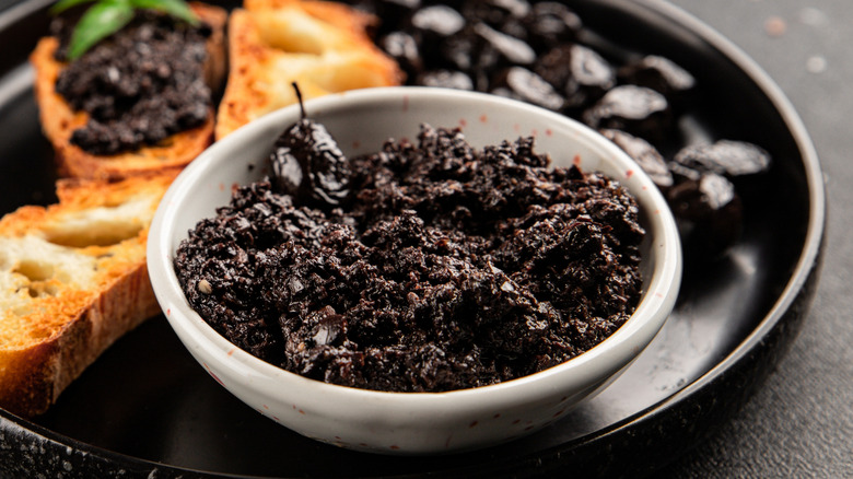 Bowl of black olive tapenade on a plate with black olives and toasted bread slices