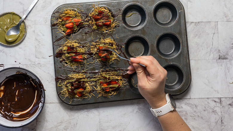 Drizzling chocolate on muffin tin with kataifi noodle nests with strawberries