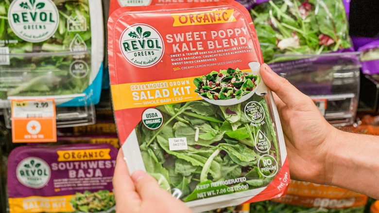 Hands holding plastic pack of Revol Greens Sweet Poppy Kale Blend greens with more Revol Greens products in background