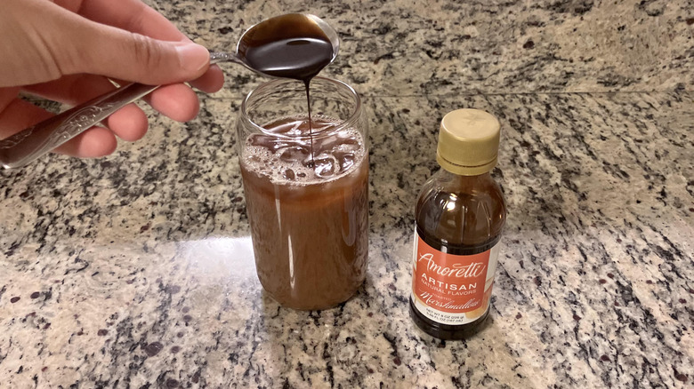 A hand pours a spoonful of Amoretti syrup into an iced coffee with a bottle of Amoretti Natural Toasted Marshmallow Artisan Flavor on the right