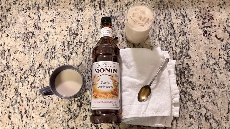 A bottle of Monin toasted marshmallow syrup sits in the center of a hot coffee, an iced coffee, and a spoonful of syrup