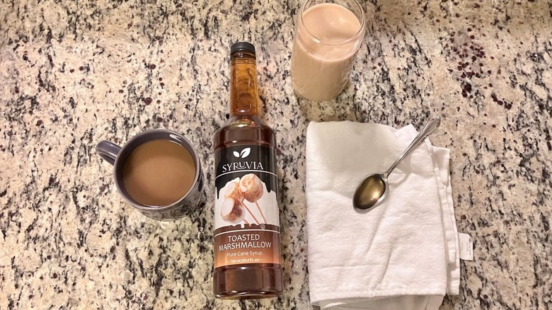 A bottle of Syruvia toasted marshmallow syrup sits in the center of a hot coffee, an iced coffee, and a spoonful of syrup