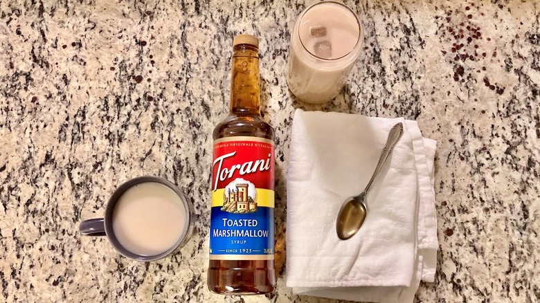 A bottle of Torani toasted marshmallow syrup sits in the center of a hot coffee, an iced coffee, and a spoonful of syrup