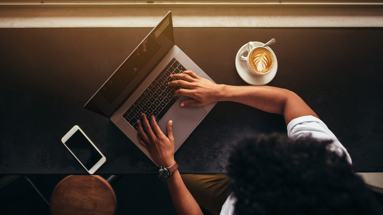 Someone working at a coffeeshop with their phone and coffee on the table next to their laptop