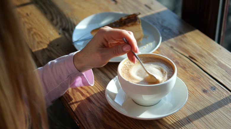Person stirs foamy coffee drink with spoon on wooden bar beside slice of cheesecake