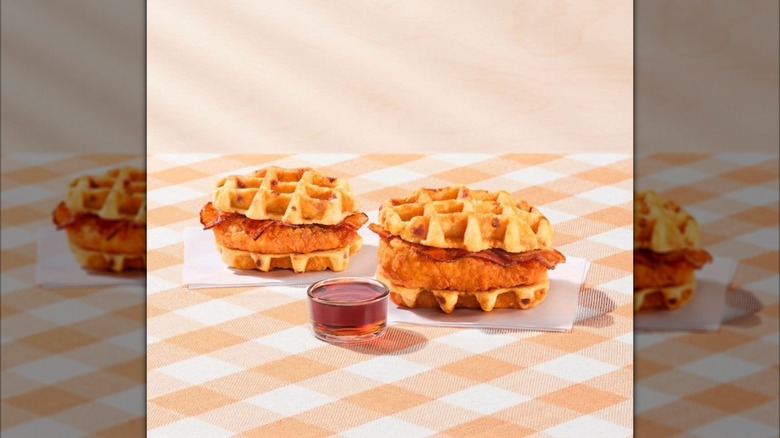 Two Chick-fil-A Chicken & Waffles Breakfast Sandwiches with bacon sitting on orange checkerboard surface next to cup of syrup