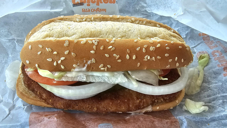 A Burger King chicken sandwich with lettuce, tomatoes, and onion spilling out the side sitting atop its paper wrapper