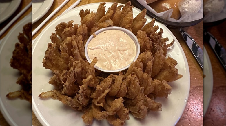 A golden fried Cactus Blossom with a cup of sauce in the center sitting on a white plate on a table