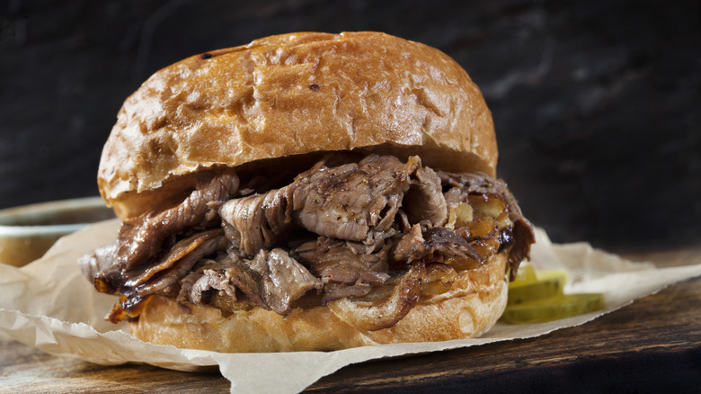 A French dip sandwich loaded with roast beef on a golden brown bun sitting on paper with Pickles in the background