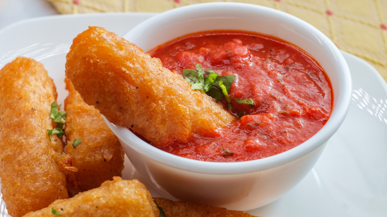 Golden crispy mozzarella sticks on a plate, one of them dunked into a small bowl of marinara sauce