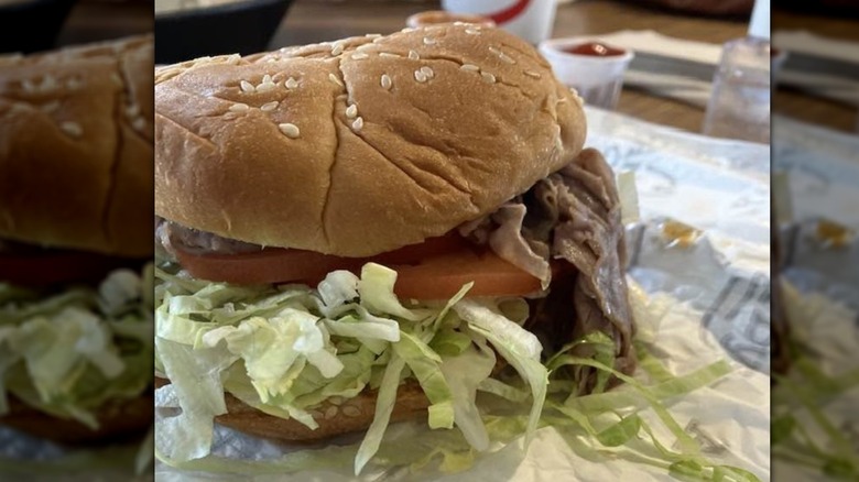 A roast beef sandwich with shredded lettuce spilling out invisible tomatoes on a sesame seed bun