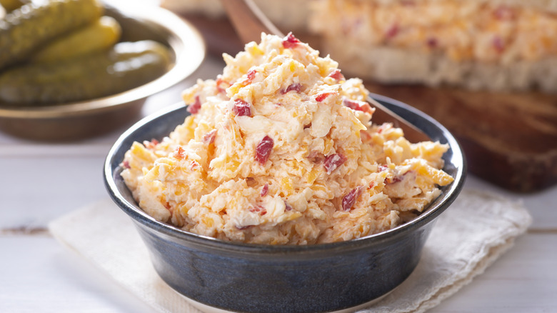 A container of pimento cheese with a bowl of pickles in the background