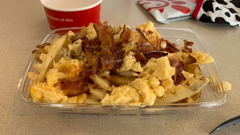 A plastic container of waffle fries topped with mac and cheese and bacon on a table.