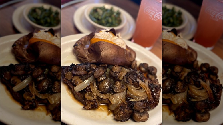 A steak topped with mushrooms and onions at Texas Roadhouse, with a baked sweet potato, green beans, and a soft drink in the background
