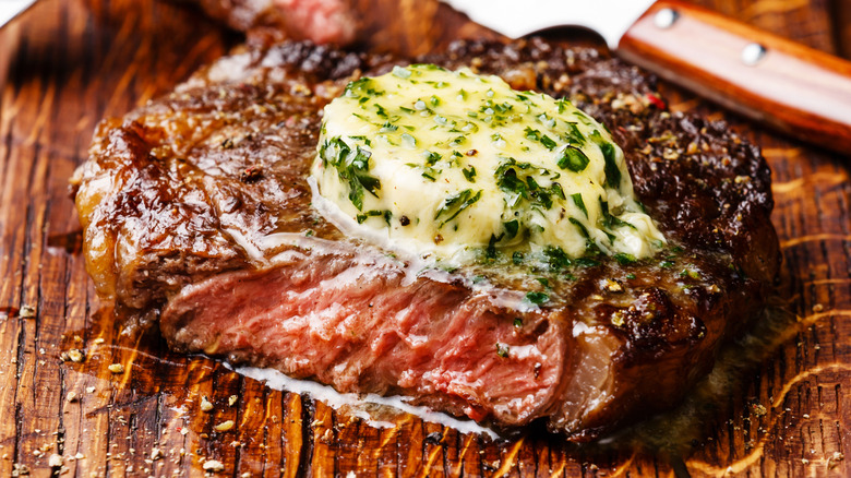 A ribeye steak topped with a pad of garlic herb butter sits on a wooden cutting board