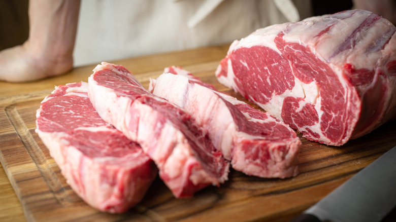 Close-up of hand-cut ribeye steaks and part of a ribeye primal on a cutting board with a butcher's hand and white apron in the background