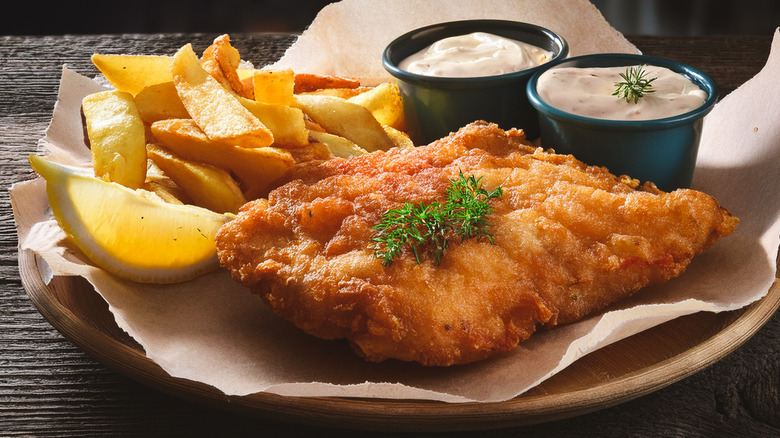 Piece of fried fish next to thick cut fries, lemon wedge, and two black cups of tartar sauce on wooden plate