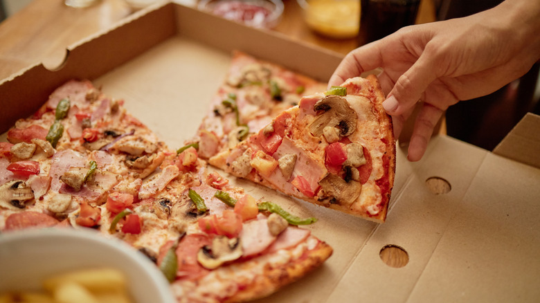 A hand picks a piece of pizza with multiple toppings on it out of a box