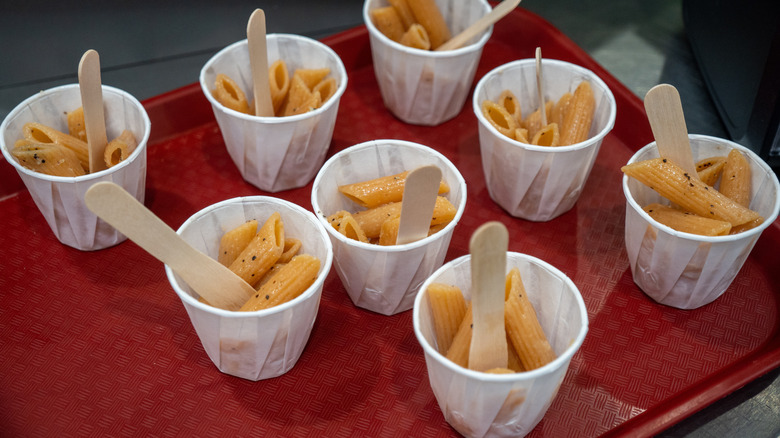 a red plastic tray filled with paper cups of cooked and seasoned penne pasta with small wooden eating utensils