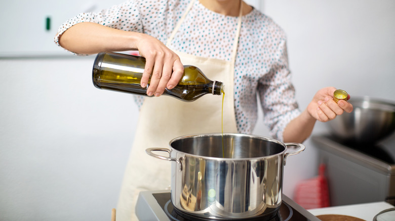 Person in a white apron pouring cooking oil from a bottle into a stockpot on the stove