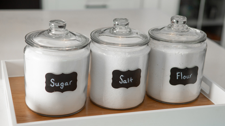 Three glass containers labeled sugar, salt, and flour sitting in a wooden container on a counter